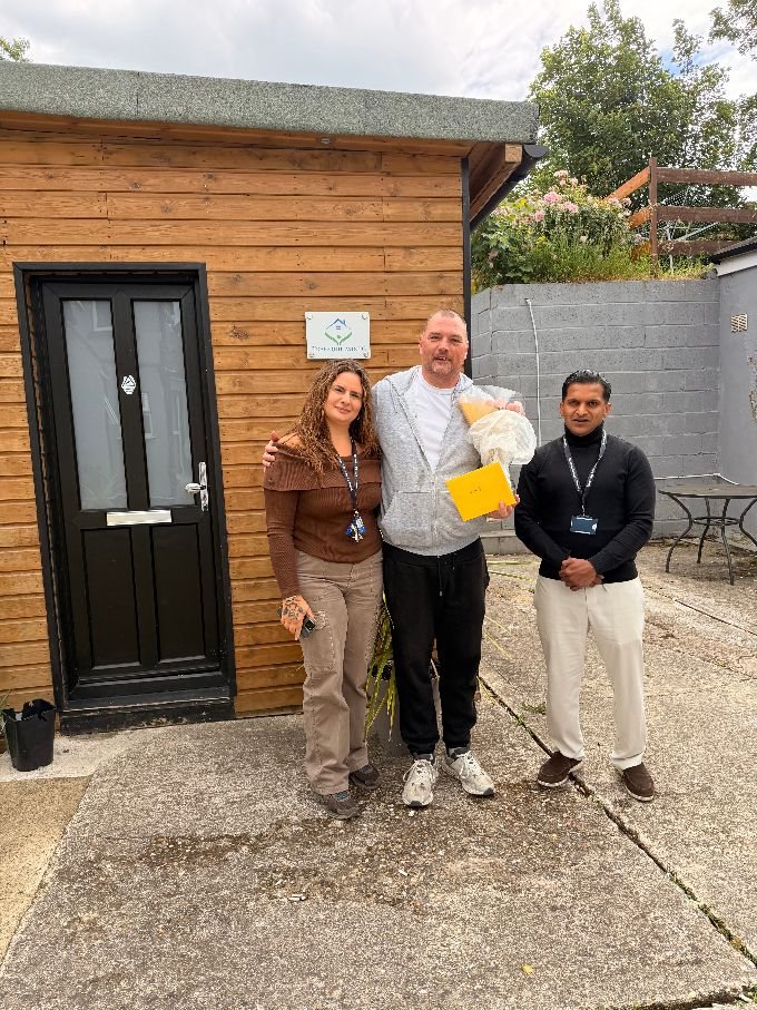 A Tranquil Minds resident standing with two support workers outside his home, holding flowers and a card to mark a milestone.